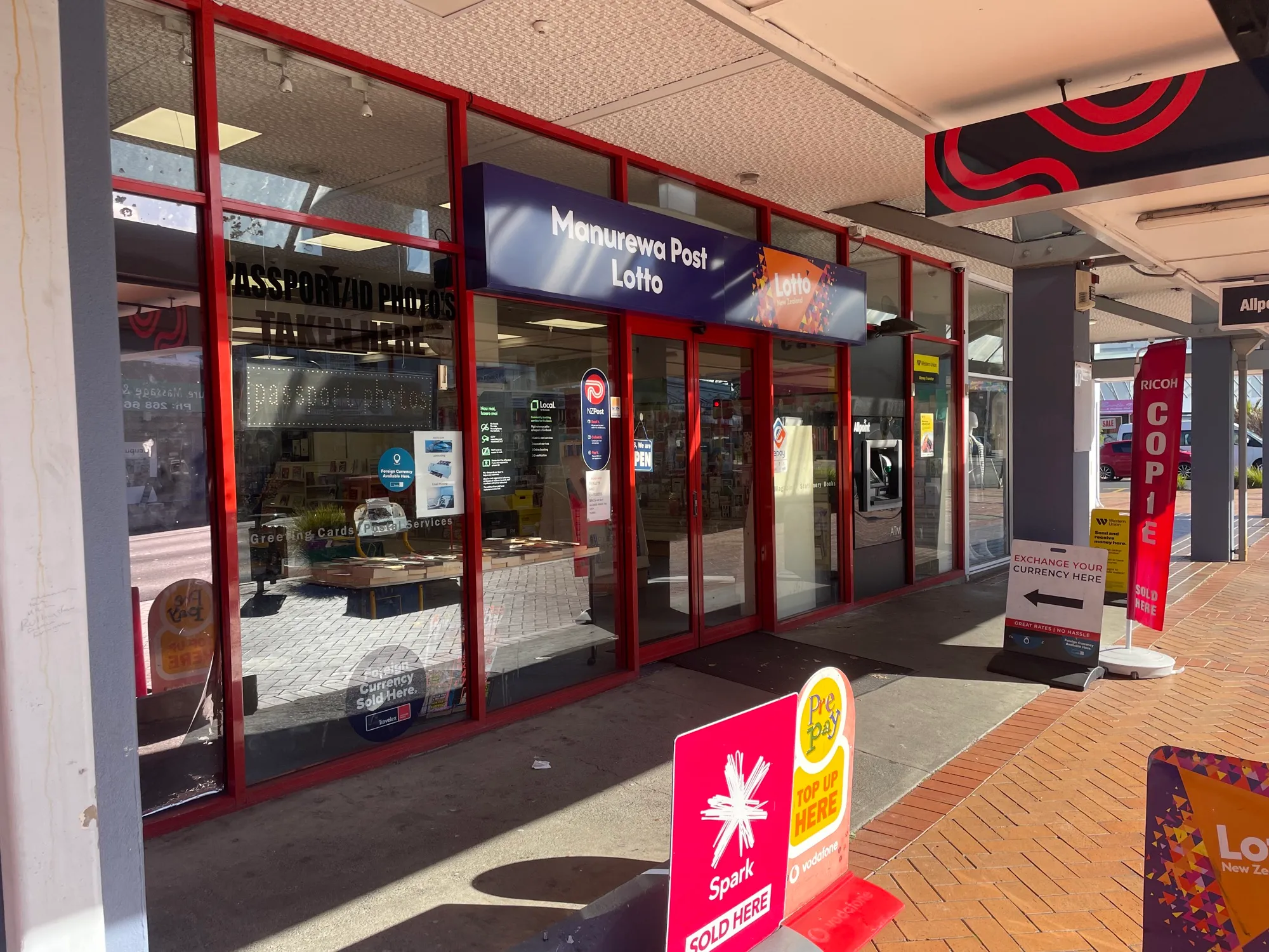 The covered walkway at Southmall, lined with shopfronts including Manurewa Post Lotto, its brick paving leading out to the sunlit entrance.
