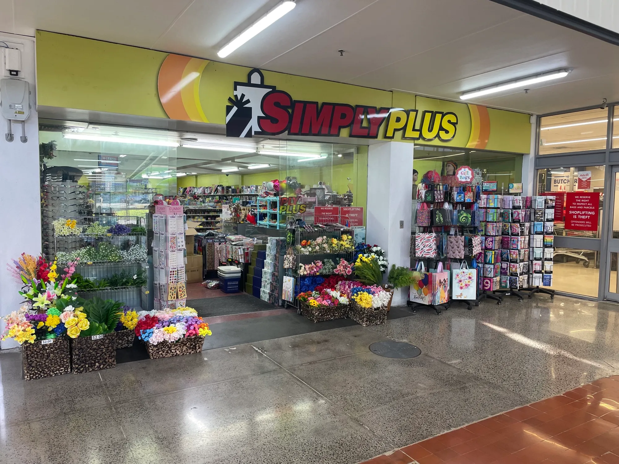 The Simply Plus shopfront opening onto Southmall's polished concourse, with buckets of artificial flowers and racks of gift bags set out across the walkway, and a butcher's shop further along.